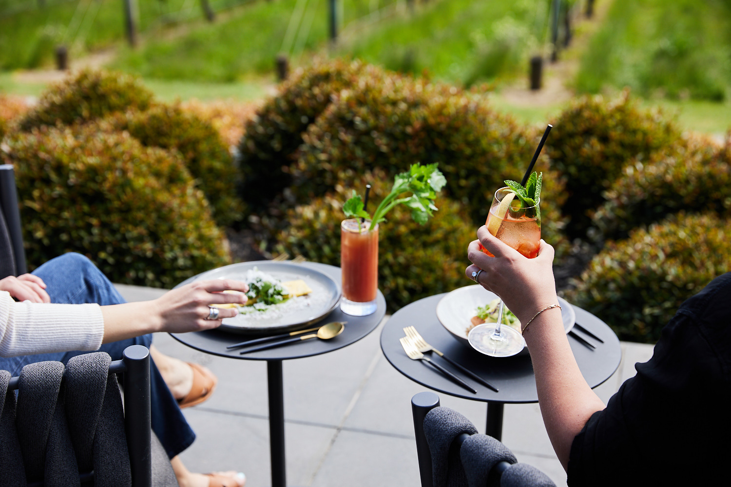 Two people enjoying cocktails and appetizers at an outdoor cafe with a blurred green natural background and tableside with drinks and plates.