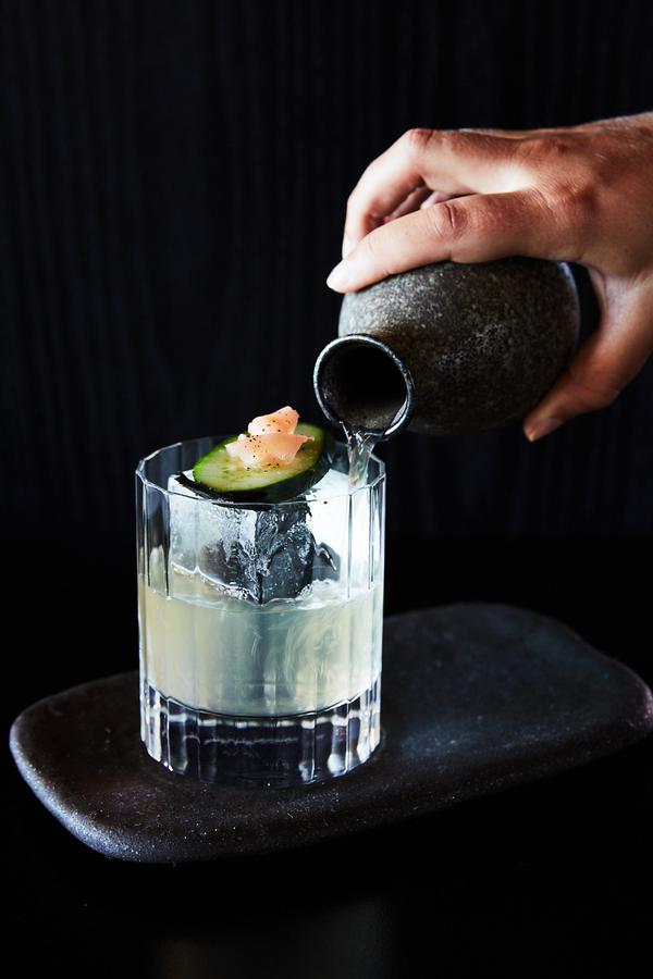 A hand pours clear liquid from a stone pitcher into a faceted glass with ice, garnished with cucumber and an orange peel, on a dark slate coaster.