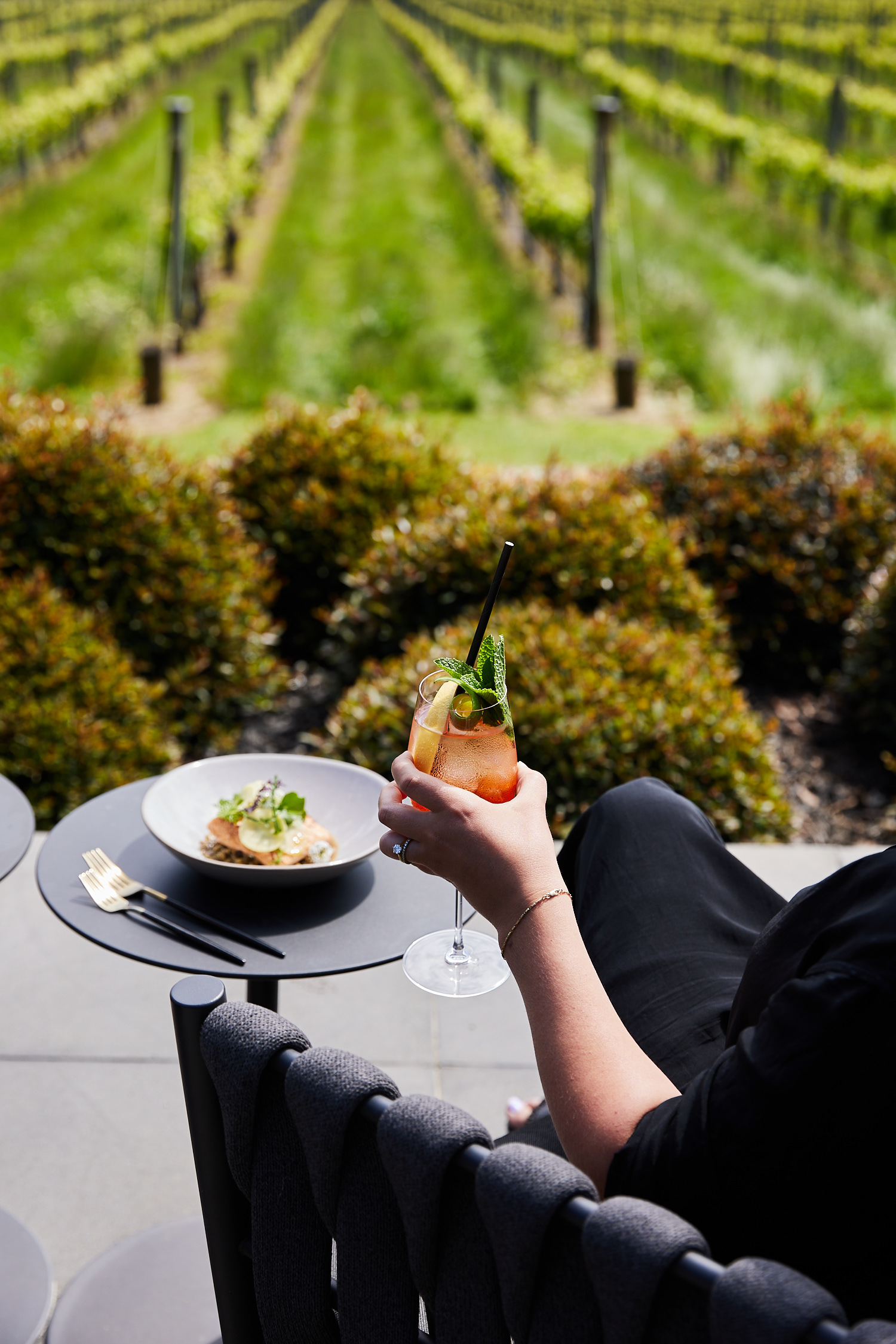 Woman enjoys a cocktail and food at an outdoor restaurant with a vineyard view in the background on a sunny day.