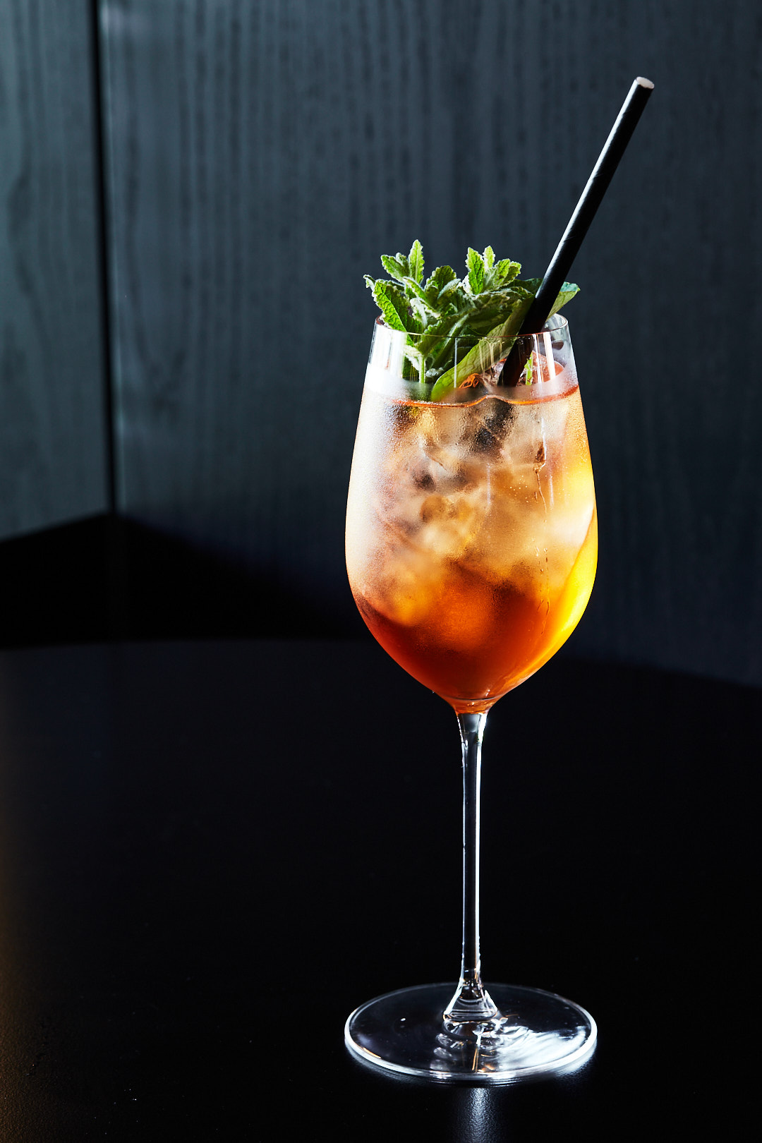 Tall glass filled with a cocktail, ice, mint sprigs, and a black straw, on a black surface with a dark wood panel background.