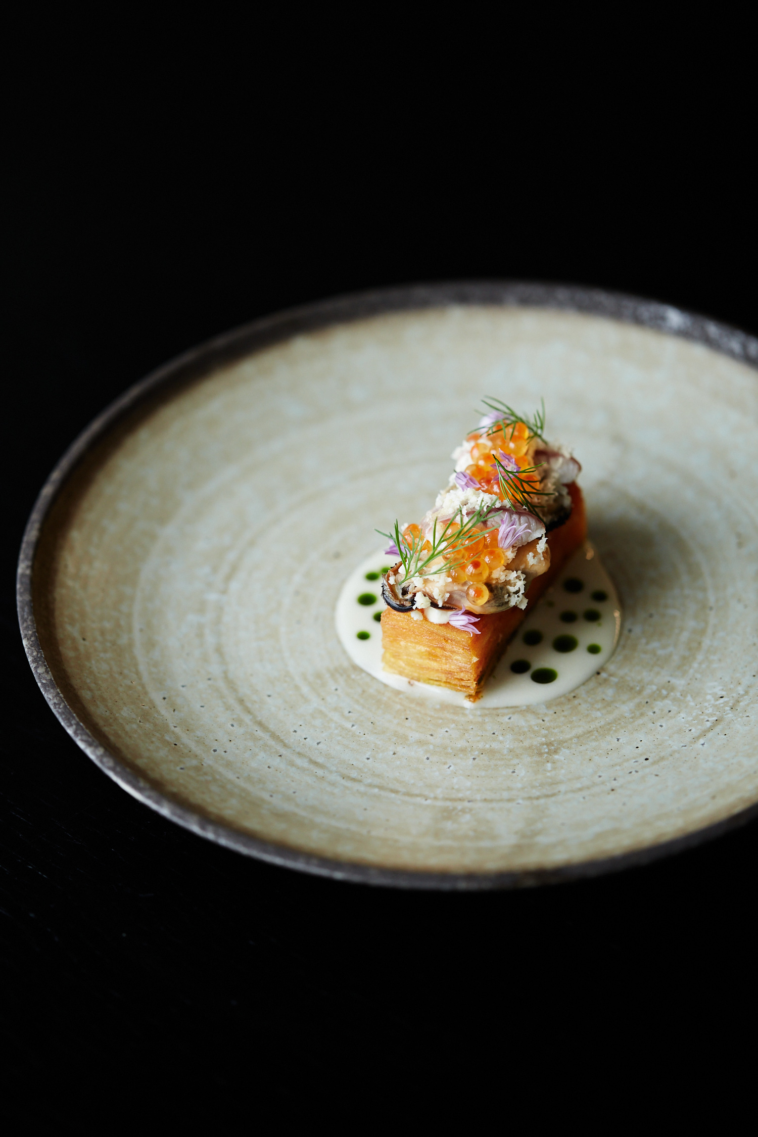 Close-up studio shot of an appetizer on a plate: stacked potatoes, creamy sauce, and orange roe, against a dark background.