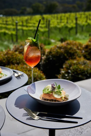 Elevated outdoor dining: A plate of garnished salmon and a tall cocktail grace a gray table, set against a backdrop of lush greenery.