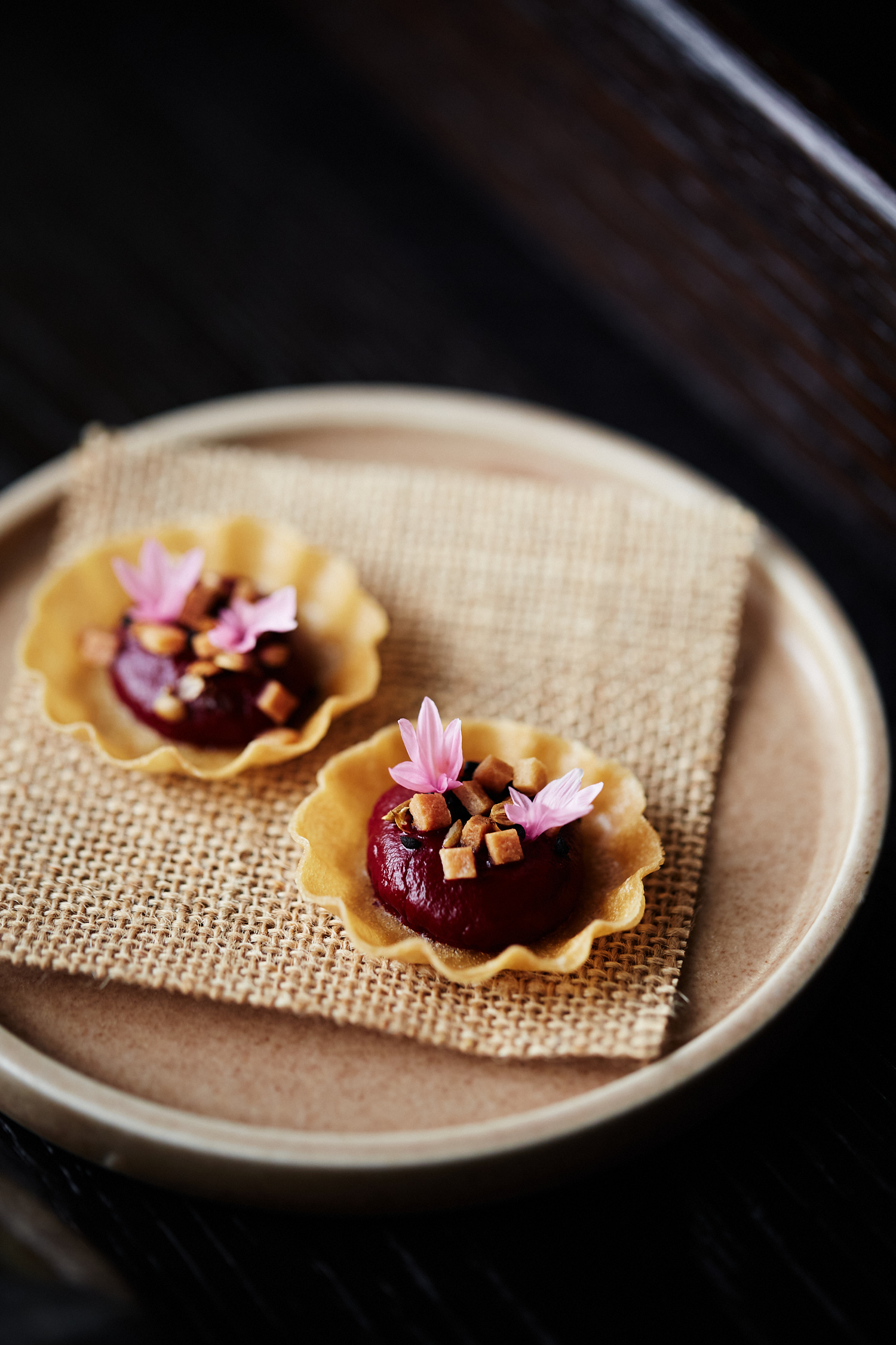 Two dessert tarts with burgundy filling are displayed on a beige plate with a textured beige mat, featuring tiny pink flowers.