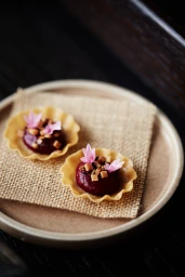 Two small tartlets with dark filling and pink flowers on a square burlap napkin resting on a round ceramic plate.