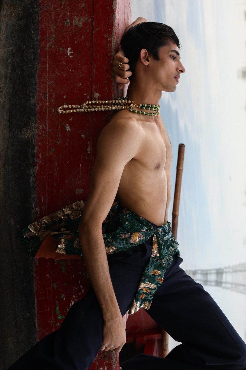 A male model poses outdoors leaning against a painted red structure. He wears dark pants, necklaces, and a tied green patterned garment.