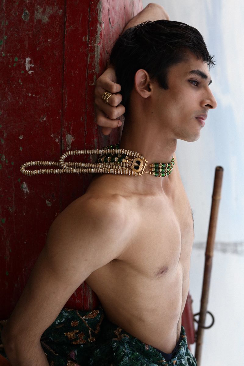 Profile portrait of shirtless adult male with green and gold beaded necklace, posing against red door, shadowed lighting.