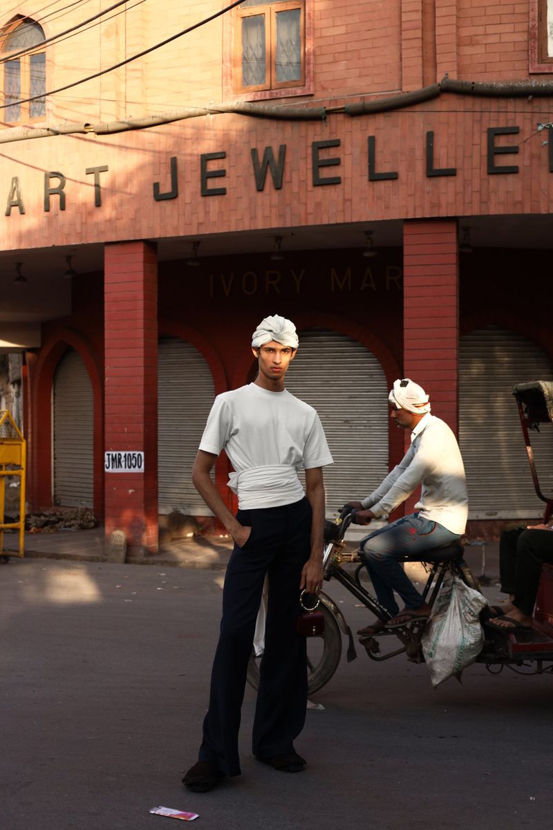 Male fashion model poses on city street in white shirt and turban, black pants, with man on bike rickshaw passing storefront sign.