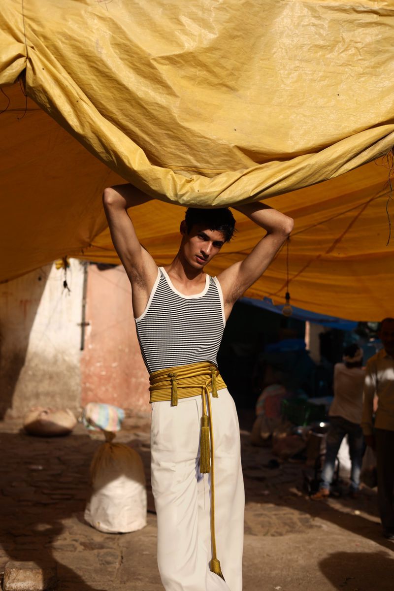 Young adult stands under a yellow tarp, wearing a striped tank top, white pants, and a gold belt, outdoors in a slightly gritty location.