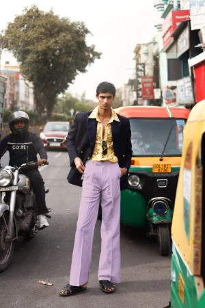 A young man poses on a busy city street wearing a navy blazer, gold shirt, and lilac trousers, with scooters and cars in the background.