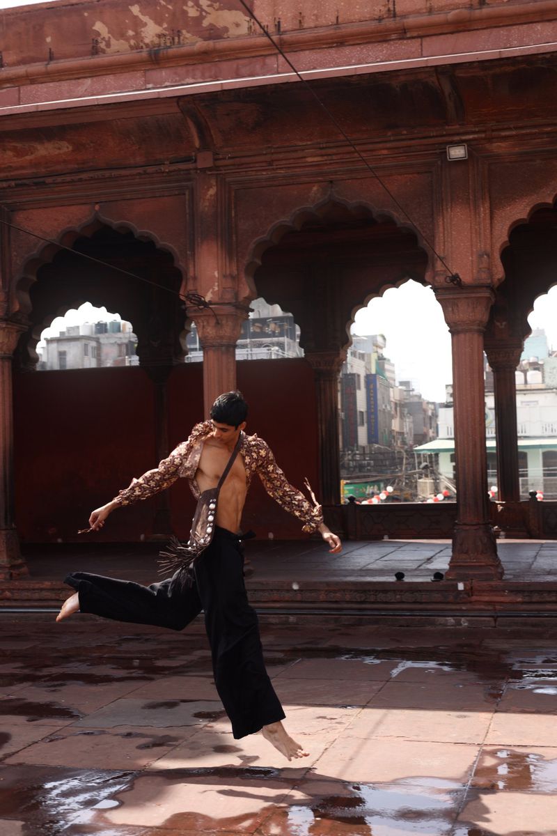 An adult male dancer jumps barefoot on a terra cotta-tiled rooftop in bright daylight with historic stone arches and a city background.