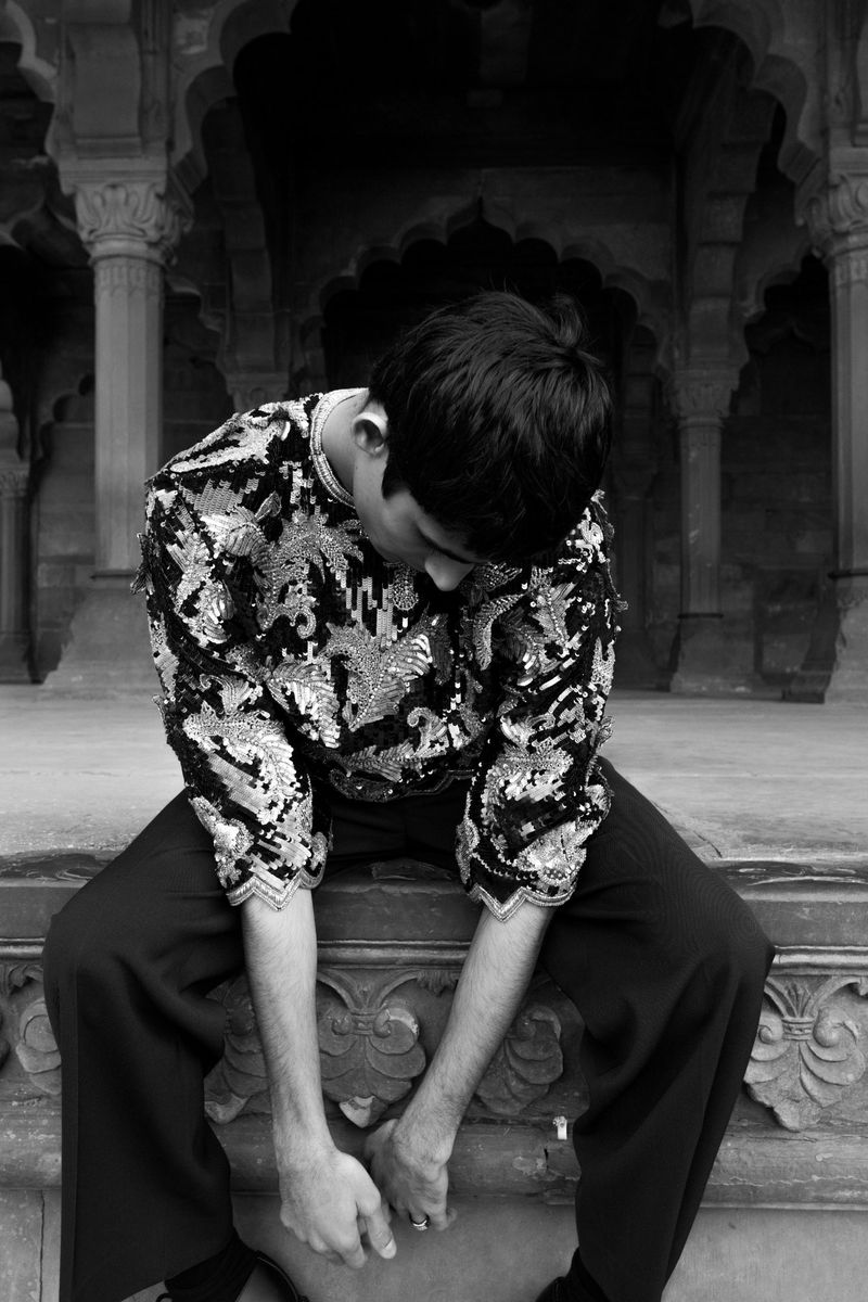Monochrome portrait of a person in a sequined top and black pants sitting on a stone ledge with arches in the background.