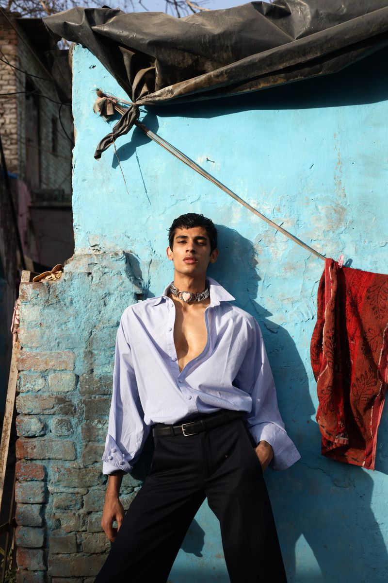 Young adult poses in an open light blue collared shirt and dark pants against a distressed wall in a sunny, outdoor setting.