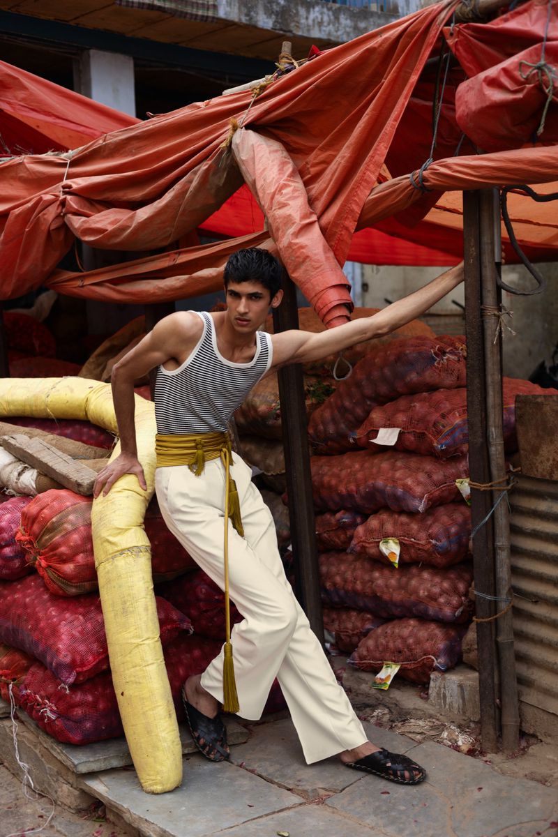 Young man poses at an outdoor market with produce, wearing a tank top and light pants with a decorative belt and tassels.