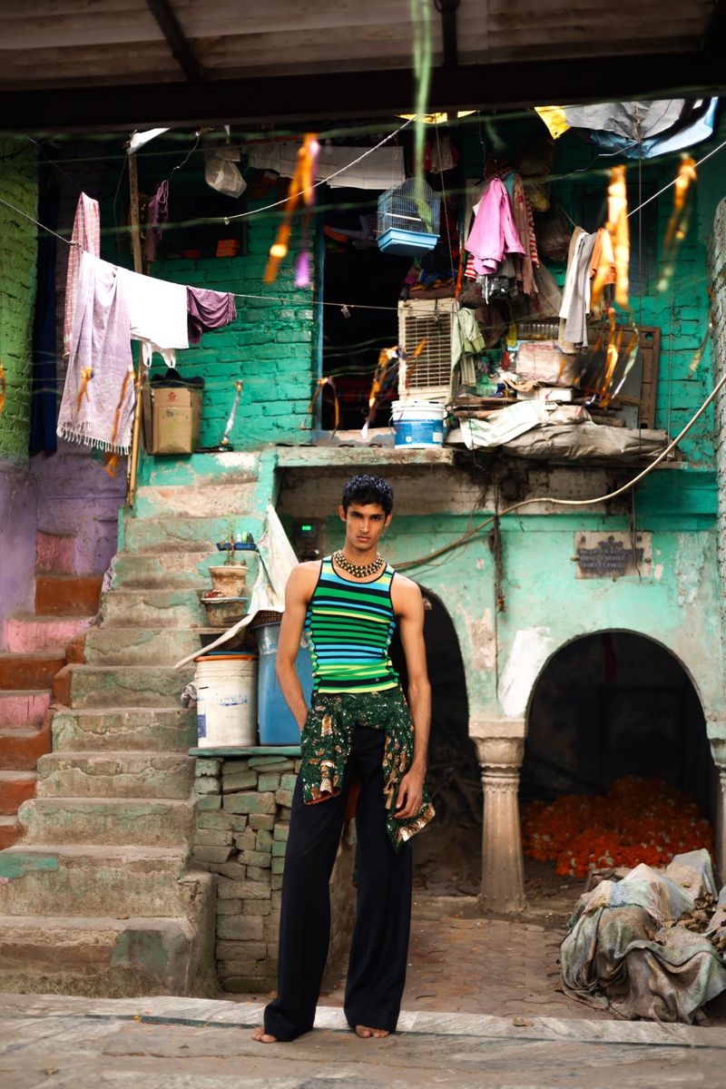 A young man poses in a striped tank top and dark pants in a courtyard setting with textured walls, a staircase, and draped clothing.