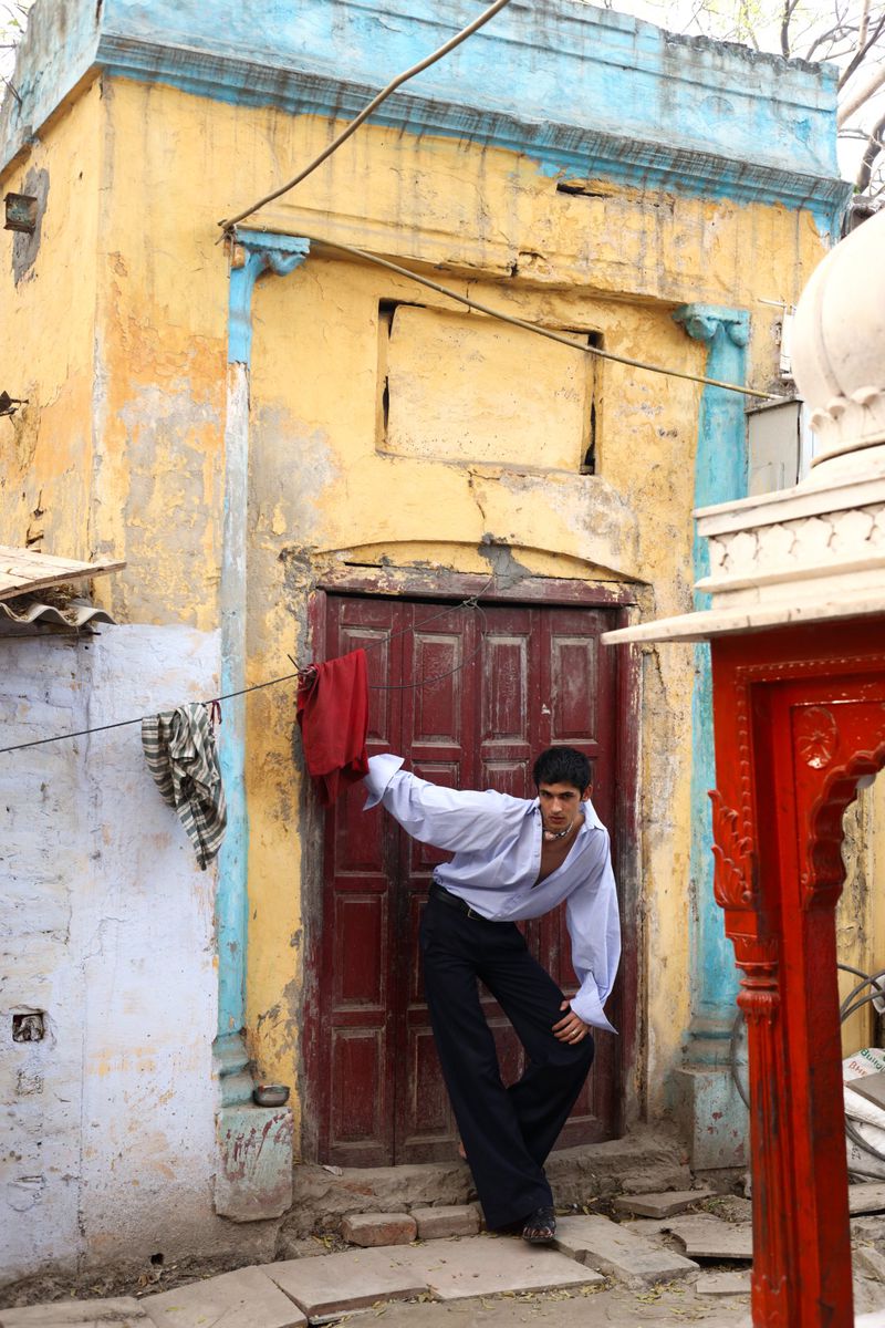 A man poses in front of a worn yellow building with red doors, wearing an unbuttoned shirt and dark pants in an urban setting.