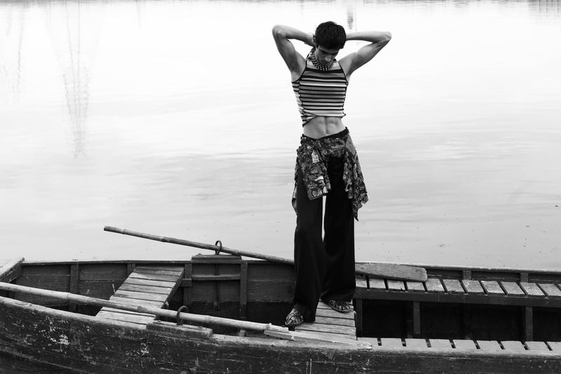 Monochrome image of a slender adult male standing on a weathered wooden boat, with hands behind his head, on a tranquil lake.