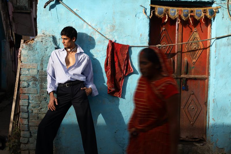 An adult male fashion model poses outdoors in a light blue collared shirt and dark dress pants in front of a light blue building.
