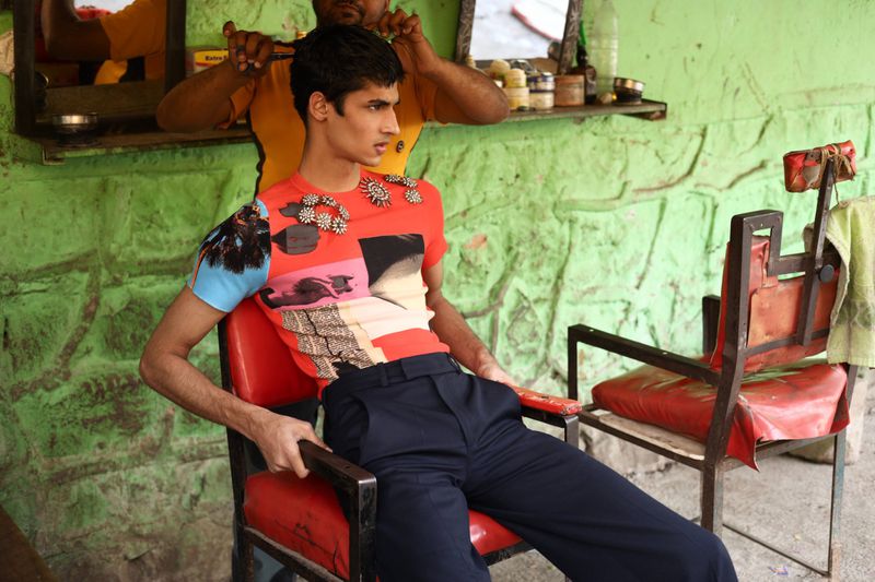 A young man sits in a red barber chair as another man cuts his hair in an outdoor barbershop with a green painted wall and miscellaneous items.