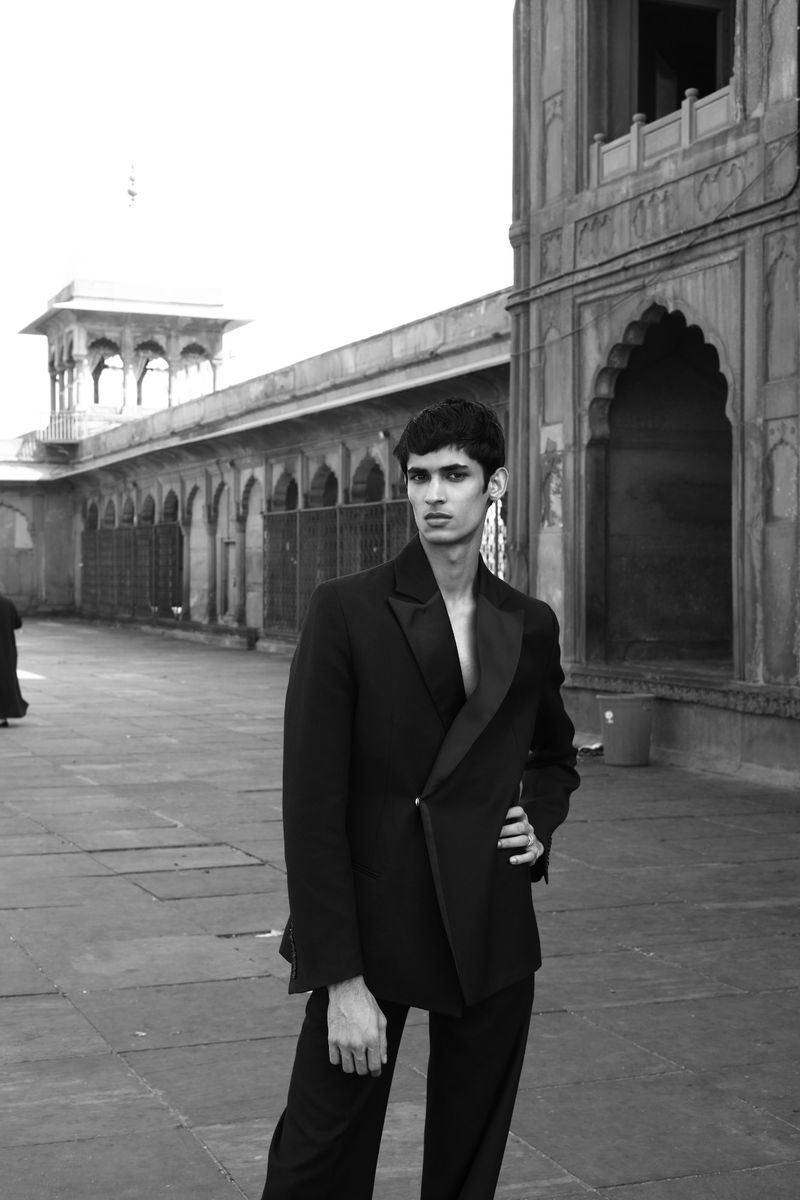 Monochrome portrait of a person in a black suit posing in front of a building with arches, showcasing fashion and architectural elements.