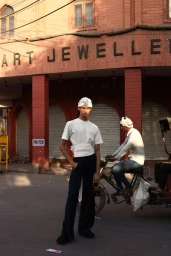 Model wearing white t-shirt and turban standing in front of a red brick storefront with a cyclist on the street