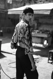 Black and white portrait of a male model wearing an ornate embroidered crop top and dark trousers outdoors in an alleyway.