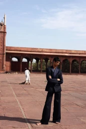 Model standing barefoot with arms crossed in a dark suit on a wide stone courtyard with a red sandstone building in the background