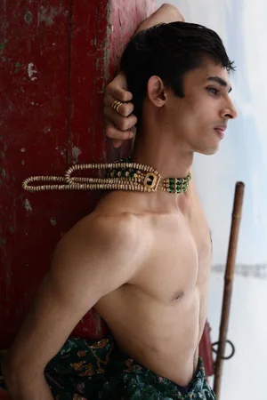 Shirtless male model wearing layered beaded necklace and gold rings, leaning against red surface