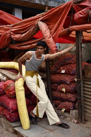 Model wearing a striped tank top and white trousers leaning against stacked sacks under an orange canopy for Les Hommes Publics Delhi
