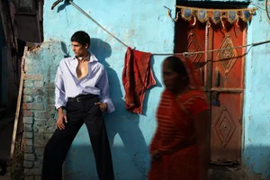South Asian man in an unbuttoned shirt leaning against a blue wall next to a woman in motion