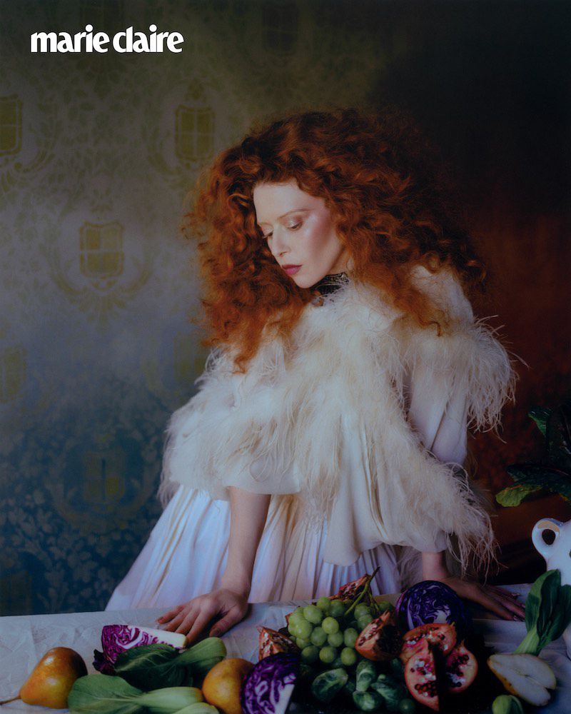 A woman with red curly hair and a white feathered dress poses behind a table with fruit in a studio with a patterned wall for Marie Claire.
