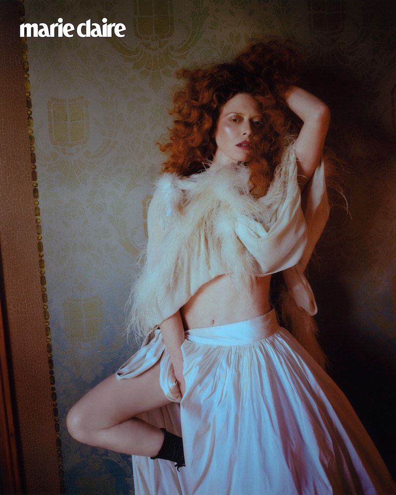 Fashion editorial shot of a woman with red hair in a white skirt and feathered shawl, posing in front of a patterned wall. 'Marie Claire' logo visible.
