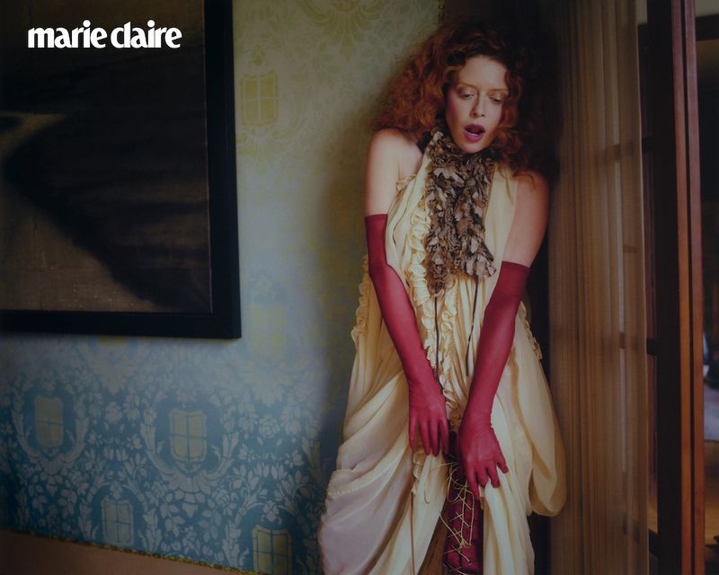 Editorial fashion shot of a red-haired model wearing a light-yellow dress and pink gloves, leaning against a wall with blue wallpaper indoors.