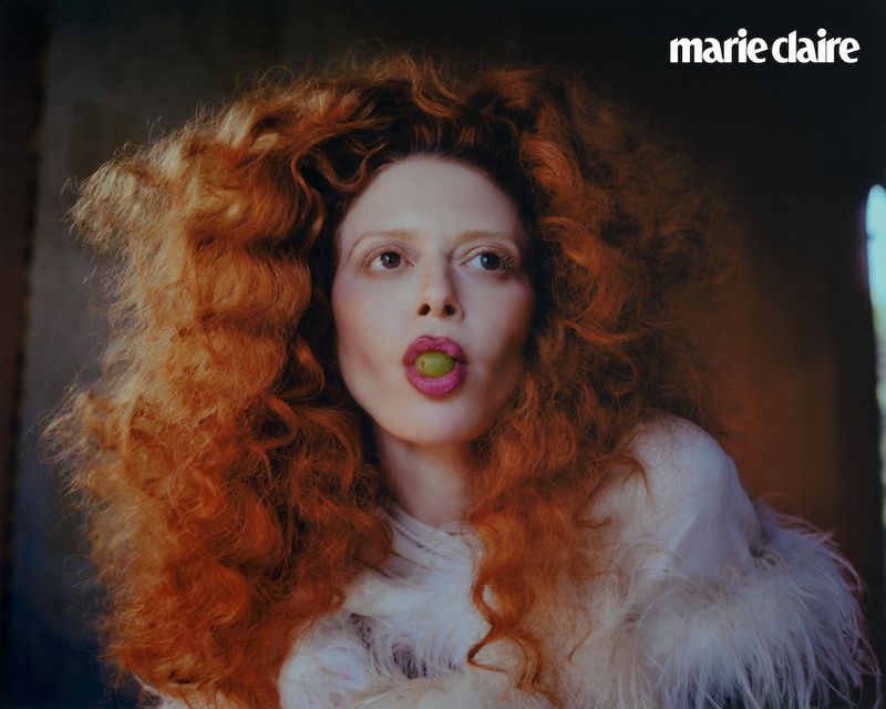 Close-up portrait of a red-haired woman with a green grape in her mouth, wearing a white, feathered garment, in warm lighting.
