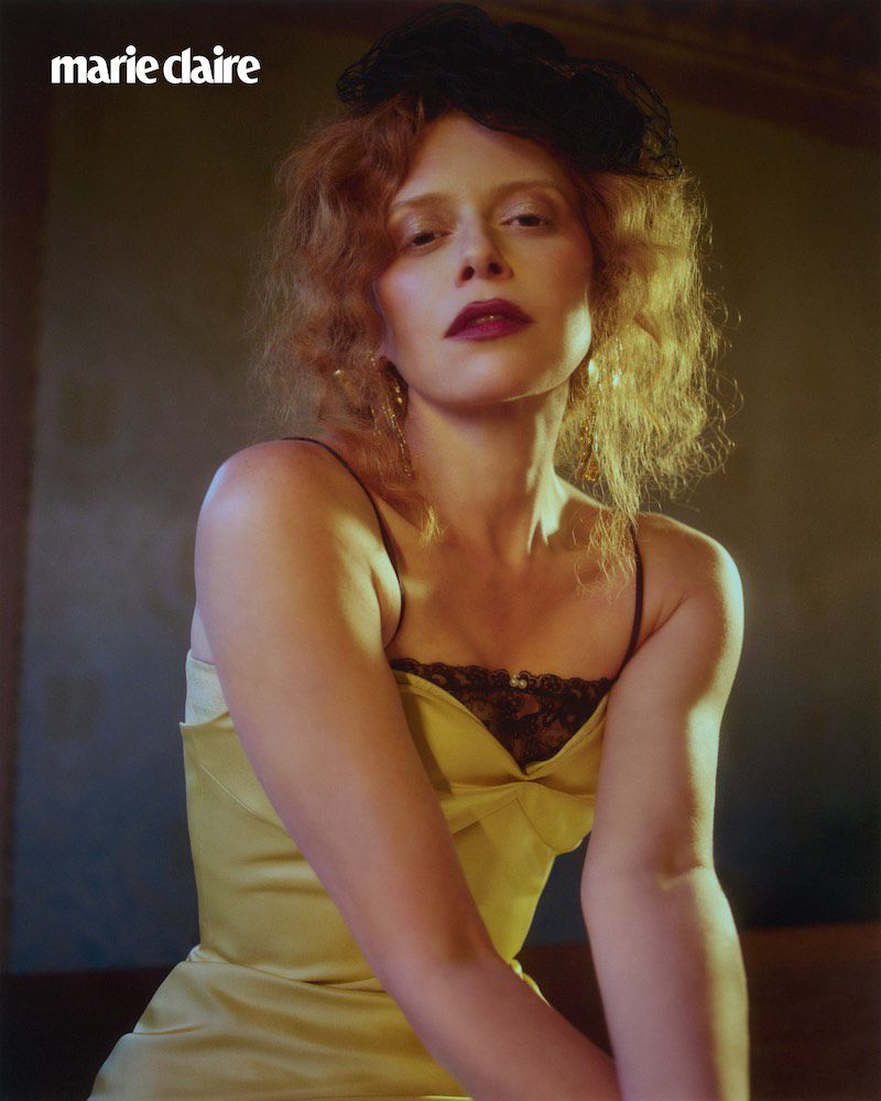 A woman with wavy red hair poses indoors in a gold slip dress; she is wearing a black fascinator and dark lipstick. The image features 'Marie Claire' logo.