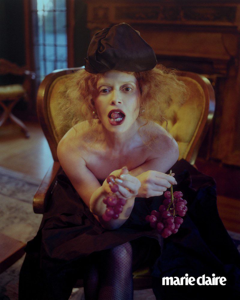 Eccentric fashion portrait of a model with auburn hair sitting in a yellow chair holding grapes indoors, wearing a black hat.
