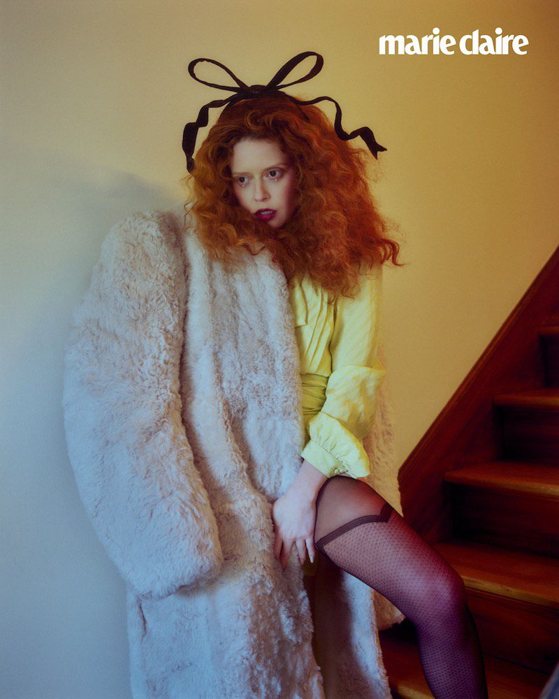 Fashion editorial shot of a woman with curly red hair wearing a yellow dress and a long white coat standing on a wooden staircase.
