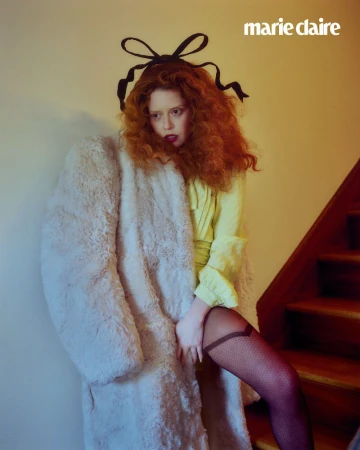 Fashion editorial shot of a woman with curly red hair wearing a yellow dress and a long white coat standing on a wooden staircase.