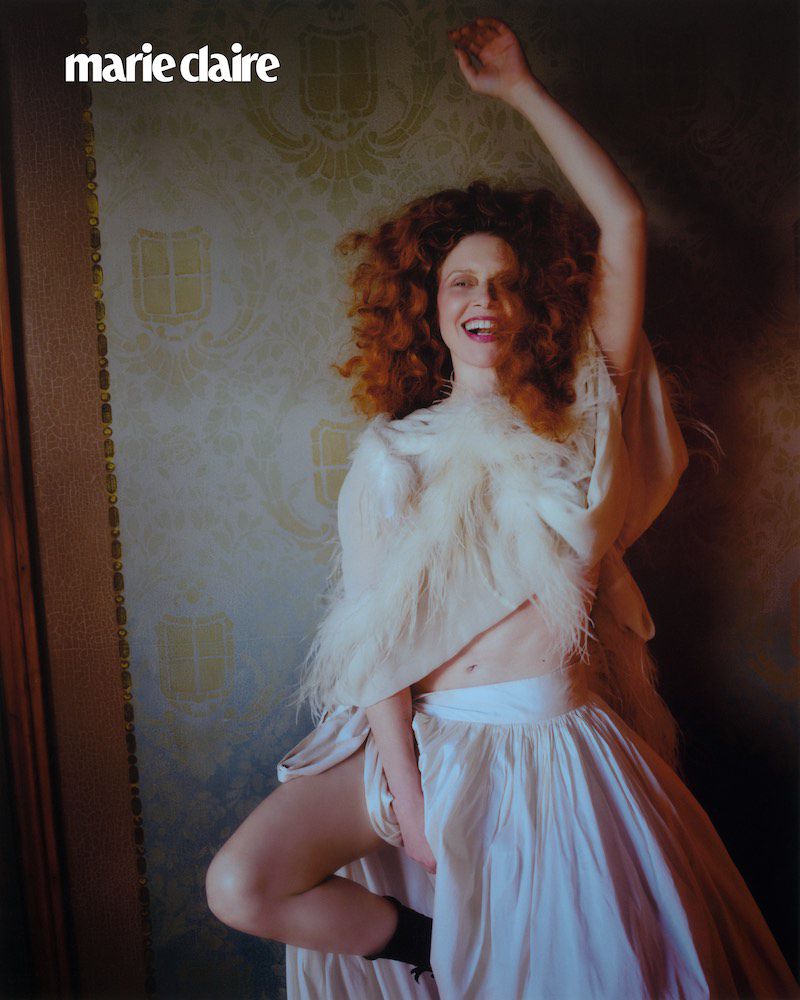 A redhead woman poses in a white dress, one leg bent, in a photograph for Marie Claire magazine against a patterned wall backdrop.