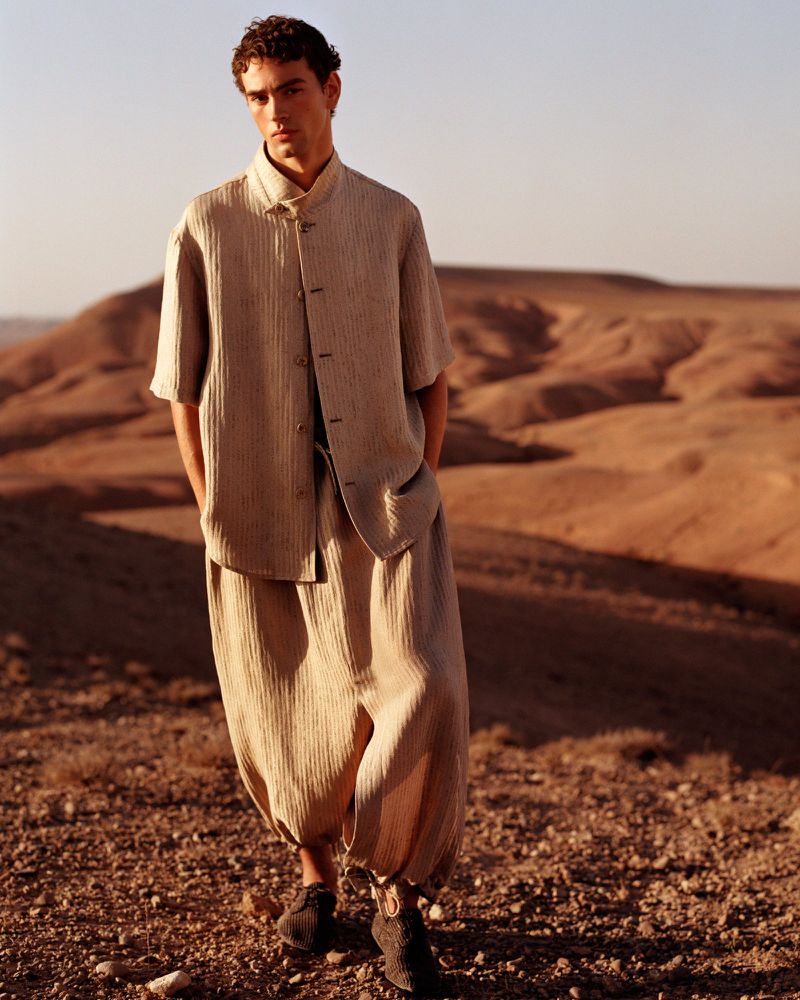 An adult male poses in loose-fitting linen clothing, featuring a short-sleeved button-up shirt and harem pants, set against a desert background.