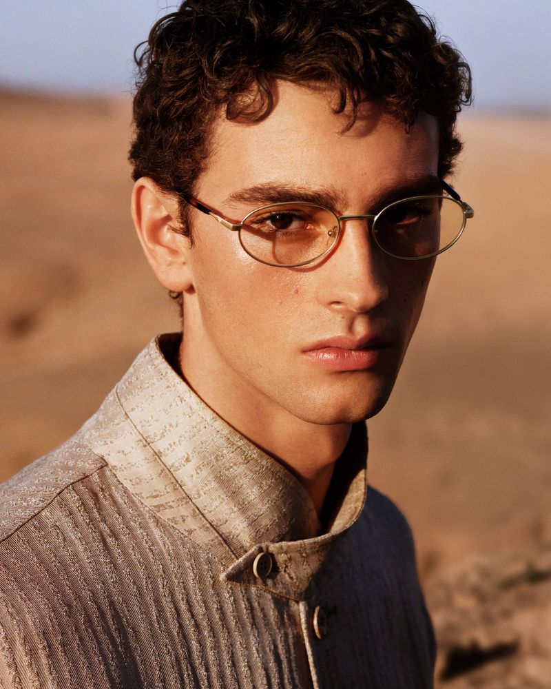 Portrait of a young adult man in vintage glasses and a tailored top, posing in a natural, desert-like outdoor environment.