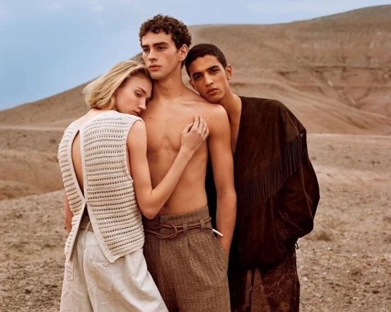 Three fashion models pose in an arid landscape, two men and one woman, in natural light, soft colour grading, styling in neutral color. 