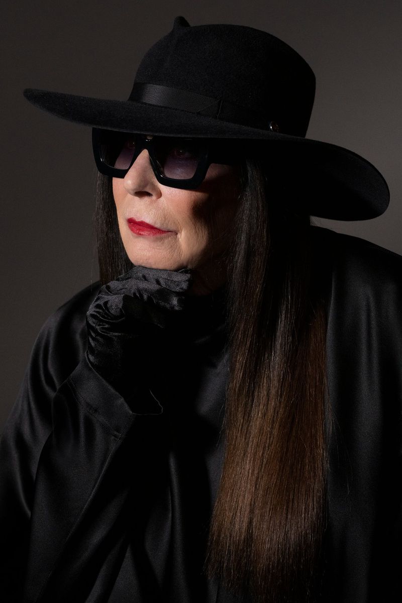 Stylish elderly woman in a black hat and sunglasses, wearing a black top, posing against a dark grey background in a studio shot.