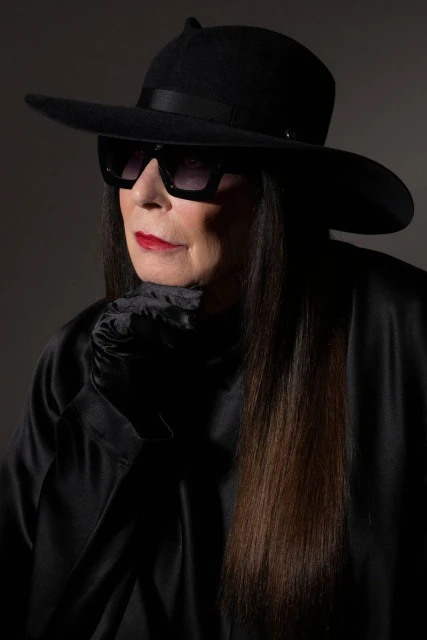 Stylish elderly woman in a black hat and sunglasses, wearing a black top, posing against a dark grey background in a studio shot.
