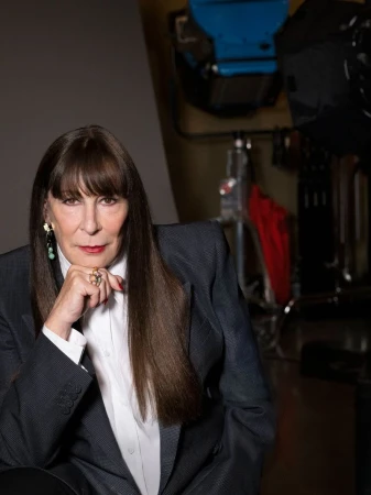 Portrait of a confident woman with long brown hair wearing a grey pinstripe blazer, posing in a photo studio with lighting equipment.