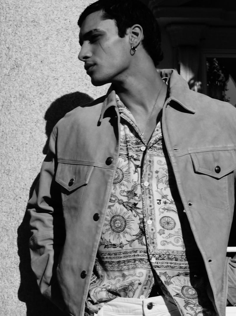 Monochrome portrait of a stylish man in a patterned shirt and jacket, standing against a textured wall with natural lighting.