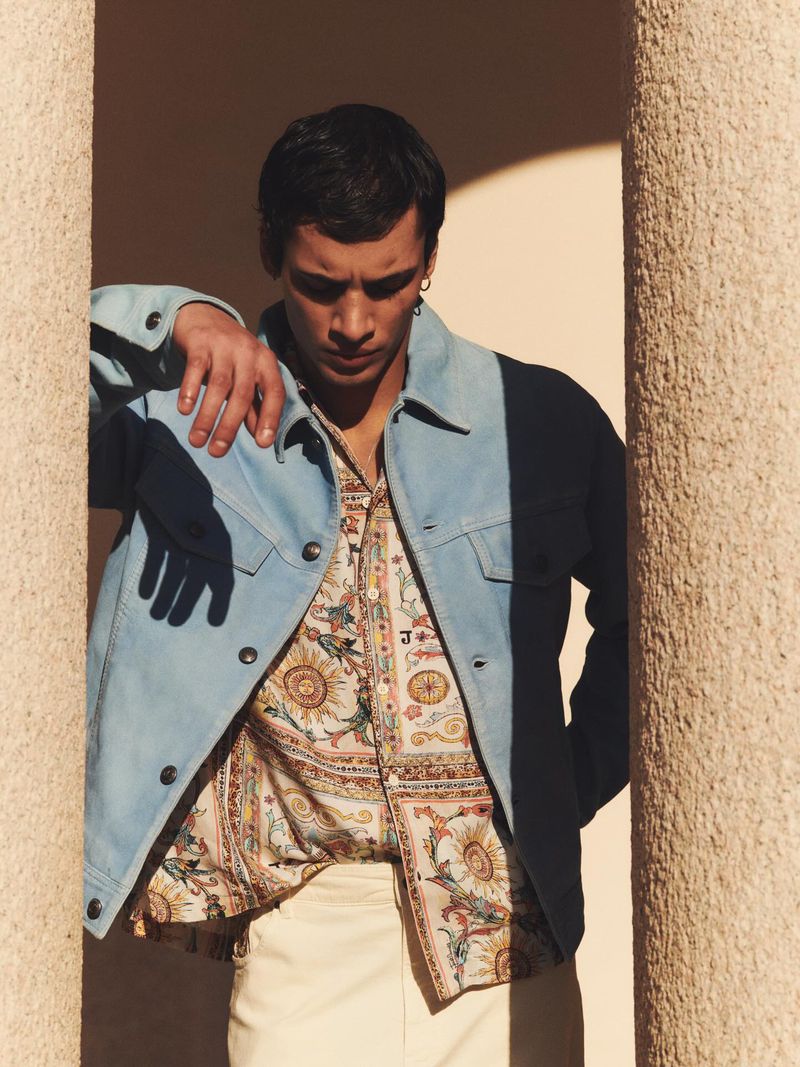 A stylish adult male poses confidently outdoors, sporting a patterned shirt, denim jacket, and white trousers next to a textured column.