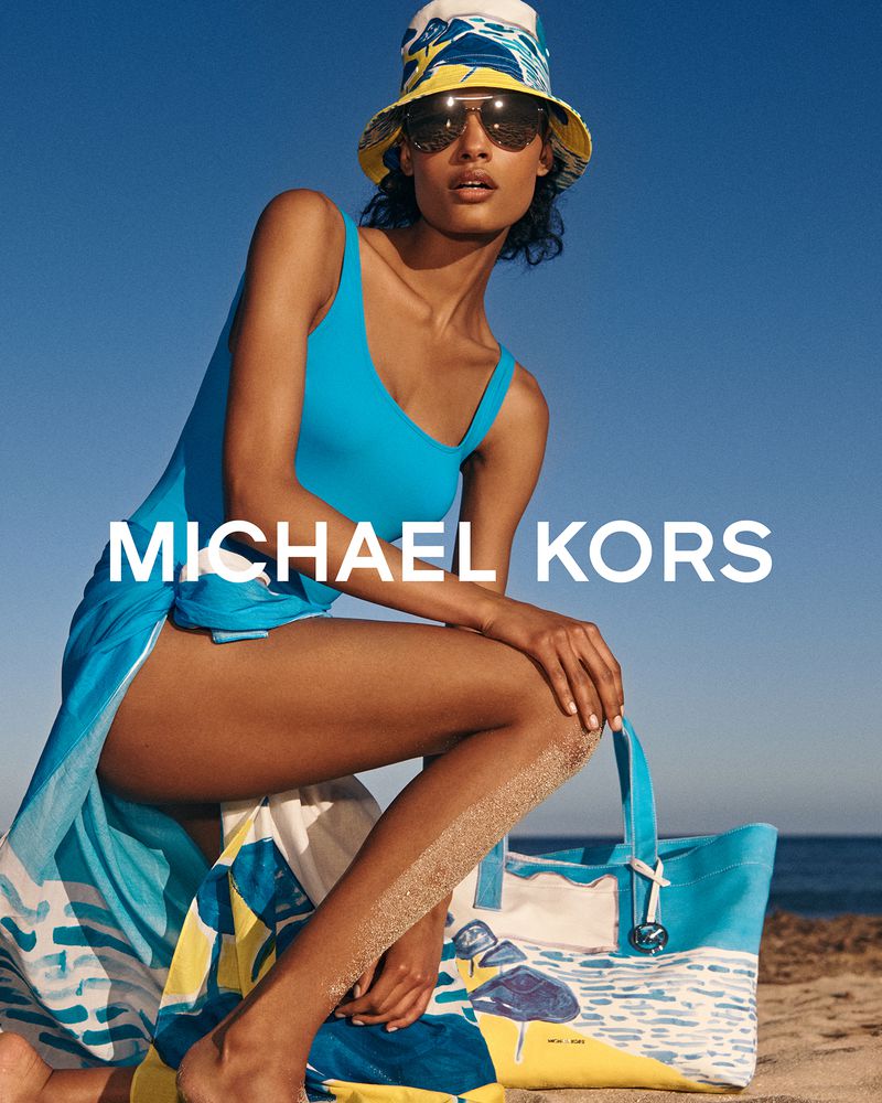 Model wearing a blue swimsuit, patterned bucket hat, and sunglasses posing on a sandy beach with a blue tote bag