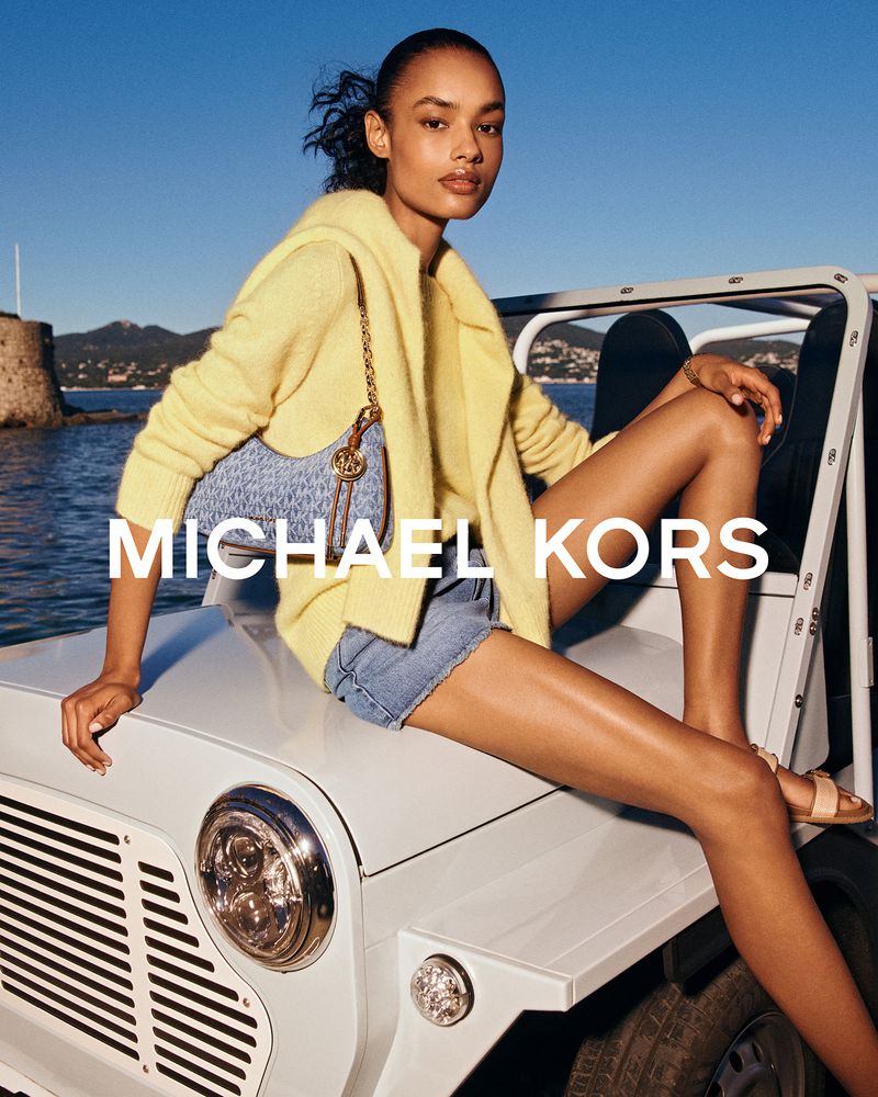 A woman sits on a white vehicle with a handbag against a clear sky and coastal backdrop, showcasing a blend of fashion and travel.