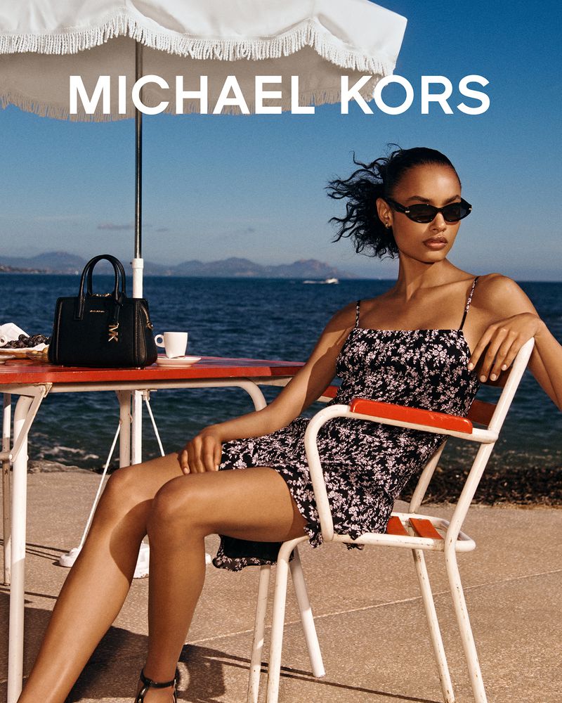 Model wearing a black floral dress and sunglasses seated at a seaside table with a black handbag under a white parasol.
