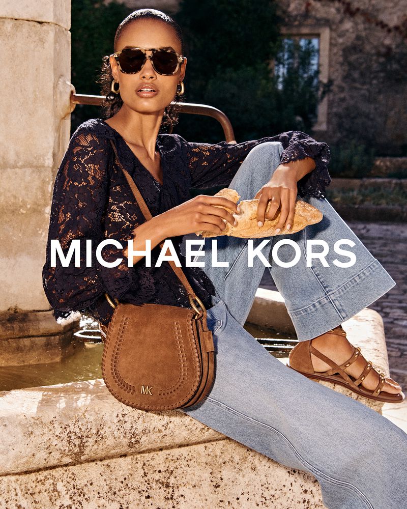 Model seated outdoors near a stone fountain wearing a black lace top, denim jeans, and holding a brown suede handbag.